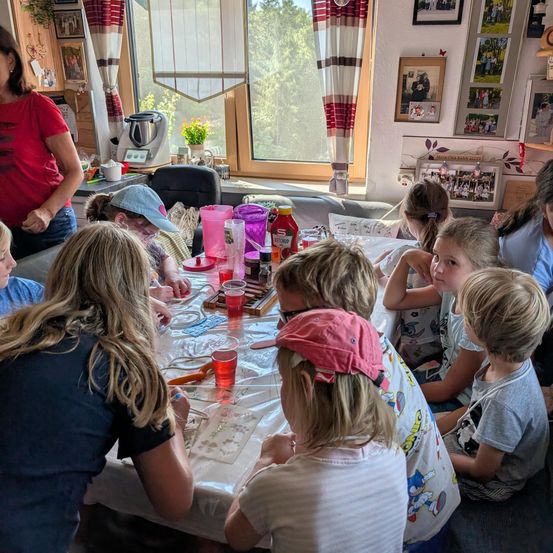 Eine Gruppe von Kindern und Erwachsenen sitzt um einen Tisch herum und ist in einer Aktivität verstrickt. Eine Frau in einem roten Shirt steht in der Nähe. Der Tisch ist mit einem weißen Tuch bedeckt und hat verschiedene Gegenstände darauf.