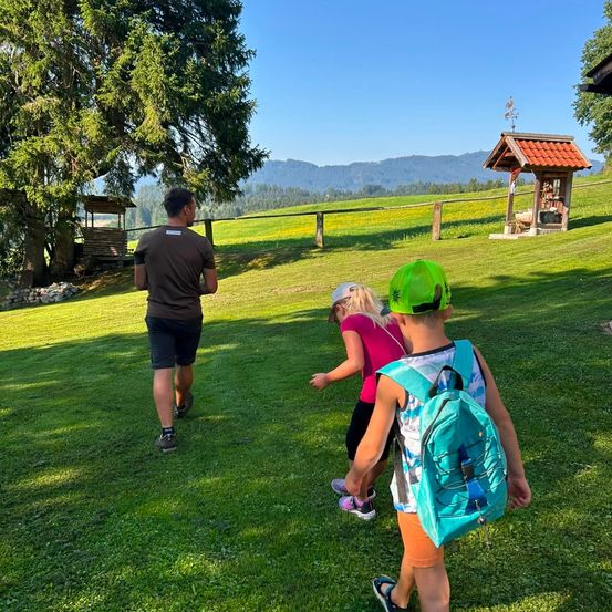 Ein Mann und zwei Kinder gehen auf einem Grasfeld mit Bergen im Hintergrund. Der Mann trägt ein braunes Shirt und Shorts, während die Kinder Mützen tragen und Rucksäcke haben.
