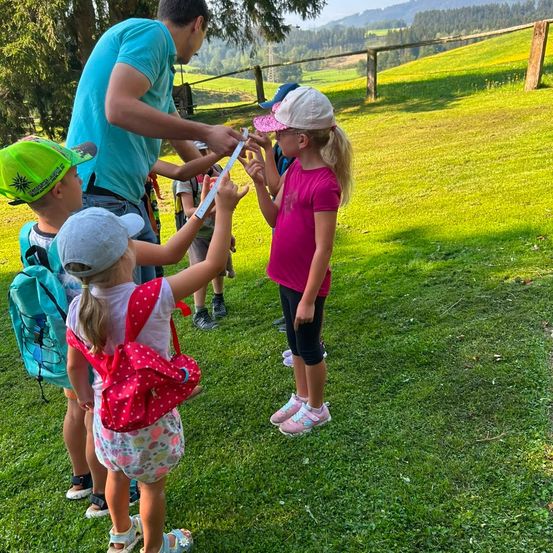 Eine Gruppe von Kindern und einem Erwachsenen steht auf einem Grasfeld, wobei der Erwachsene die Größe eines der Kinder mit einem Messstab misst.