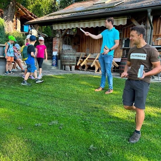 Eine Gruppe von Kindern und zwei Erwachsenen steht auf einem Rasen vor einem Holzhaus. Die Erwachsenen halten einen Stock und ein Buch, und die Kinder tragen Hüte und Turnschuhe.