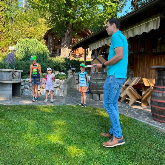 Ein Mann, der ein Buch und einen Stift in der Hand hält, steht vor drei Kindern in einem Garten. Die Kinder tragen Hüte und Turnschuhe. Dahinter steht ein Haus mit einem Holzfass und einem Holztisch.