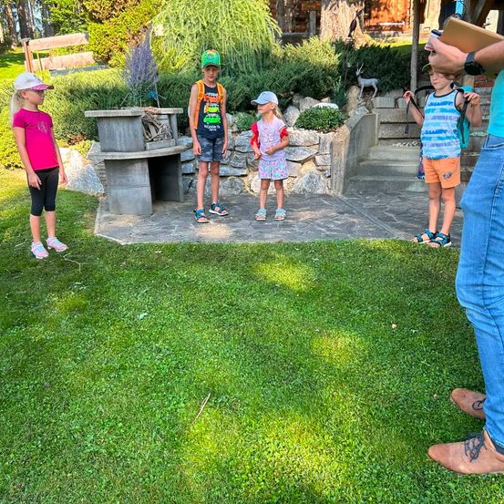 Eine Gruppe von Kindern und einem Erwachsenen steht auf einer Betonterrasse mit einer Steinmauer im Hintergrund. Die Kinder tragen Freizeitkleidung und einige haben Mützen. Der Erwachsene hält eine Clipboard.