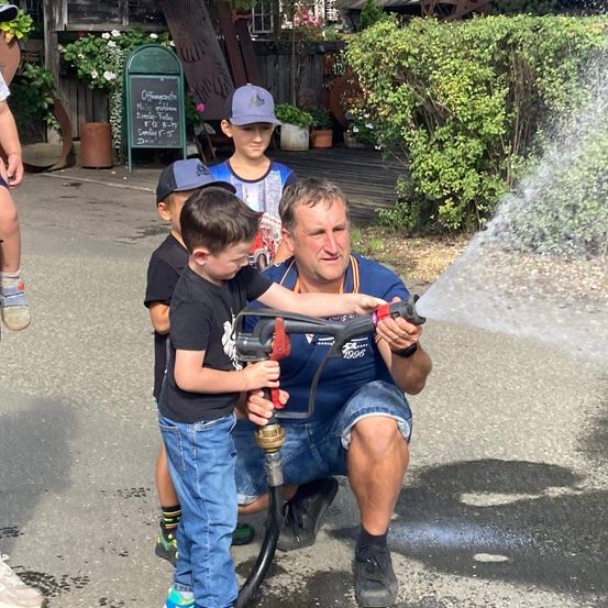 Ein Mann und drei Kinder spielen an einem sonnigen Tag mit einem Wasserschlauch. Der Mann kniet und hält den Schlauch, während die Kinder stehen und zusehen.