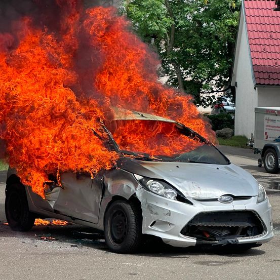 Ein silberner Ford-Wagen, der von Flammen umhüllt ist, auf einer Straße mit einem Haus und Bäumen im Hintergrund.