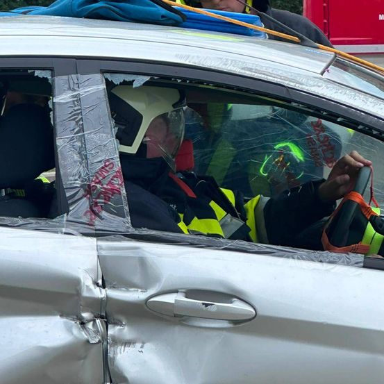 Eine Person mit Helm und Warnweste sitzt in einem beschädigten Auto, mit Rettungskräften in der Nähe.