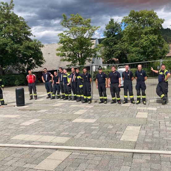 Eine Gruppe Feuerwehrleute in Uniform steht vor einem Gebäude mit Bäumen und einem bewölkten Himmel im Hintergrund.