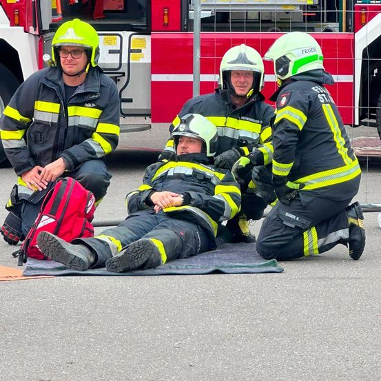 Vier Feuerwehrleute in gelben und schwarzen Uniformen, einer liegt auf dem Boden, zwei knien und einer sitzt. Eine rote Feuerlöschtasche ist neben dem sitzenden Feuerwehrmann. Ein roter Feuerwehrwagen ist im Hintergrund.
