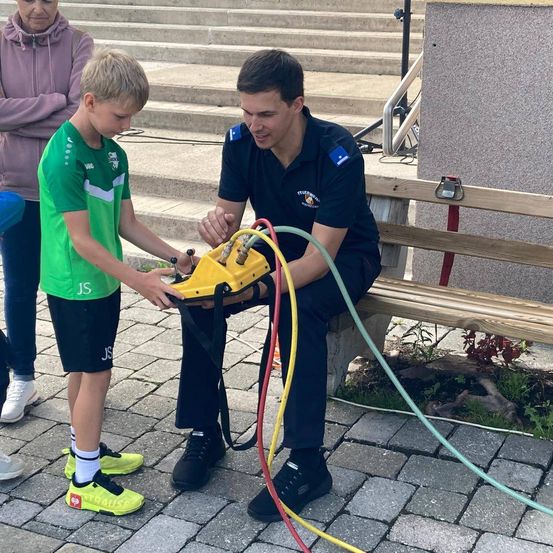 Ein Mann in Uniform sitzt auf einer Bank und hilft einem jungen Jungen, ein gelbes Gerät einzustellen. Der Junge trägt ein grünes Shirt und Turnschuhe. Hinter ihnen steht eine Frau in der Nähe einiger Stufen.