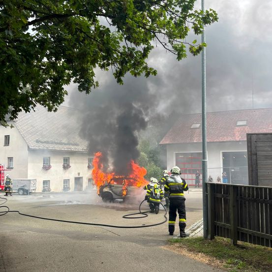 Feuerwehrleute löschen ein brennendes Auto vor einem Haus mit rotem Dach und Zaun.