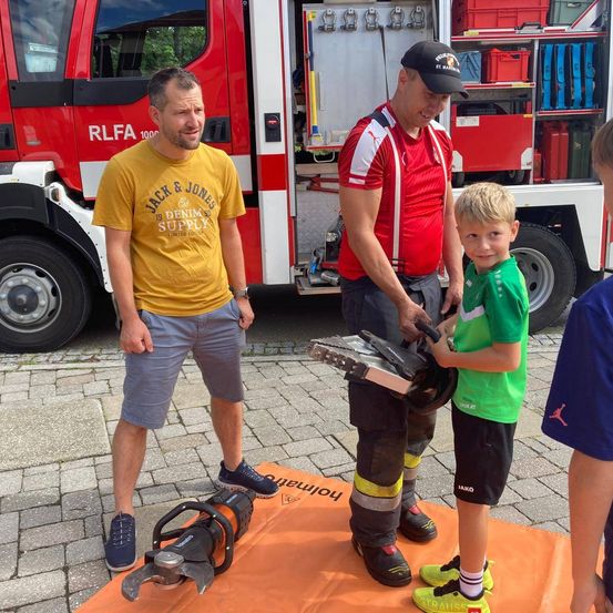 Ein Feuerwehrmann zeigt einem kleinen Jungen, wie man eine Kettensäge benutzt, während er vor einem Feuerwehrwagen steht.