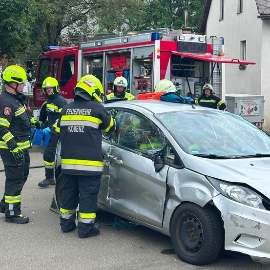 Feuerwehrleute in Schutzkleidung stehen um ein beschädigtes Auto, einer benutzt ein Werkzeug am Fahrzeug. Ein Feuerwehrwagen ist in der Nähe geparkt.
