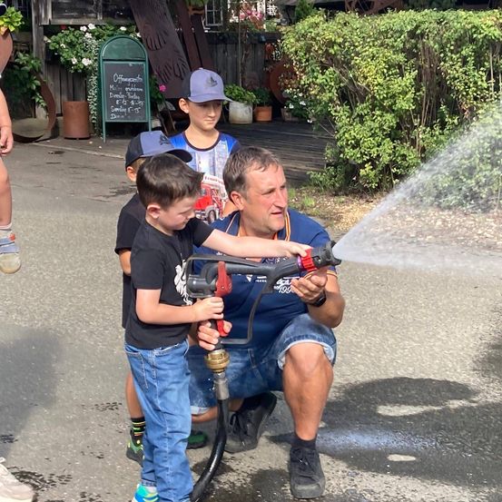 Ein Mann kniet auf dem Boden und hält einen Wassersprüher, während zwei Jungen neben ihm stehen.