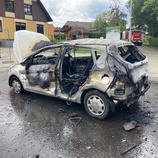 Ein ausgebranntes Auto steht am Straßenrand, mit offenem Motorhaube und zerbrochenen Fenstern. Das Innere ist verkohlt, und Trümmerteile sind verstreut.
