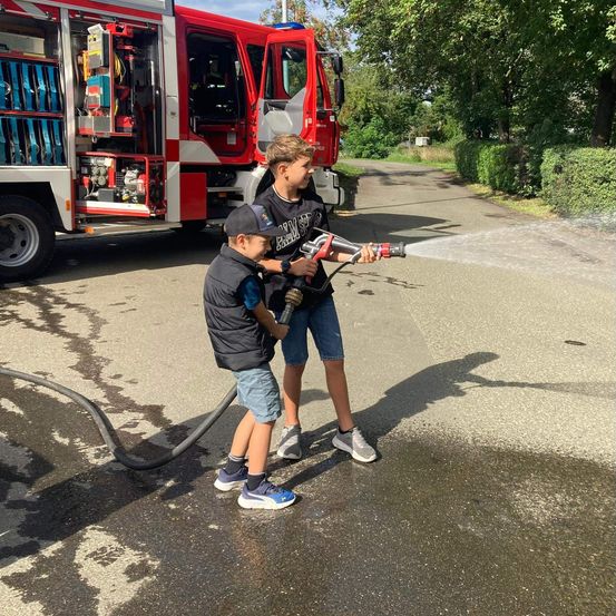 Zwei Kinder sprühen Wasser aus einem Feuerwehrschlauch auf eine nasse Straße. Ein roter Feuerwehrwagen ist in der Nähe geparkt.