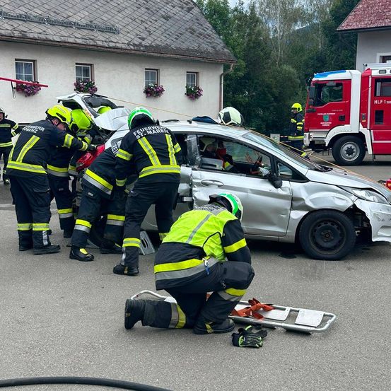 Feuerwehrleute retten eine Person aus einem beschädigten Auto. Sie tragen Helme und Uniformen. Ein Feuerwehrwagen ist in der Nähe geparkt.