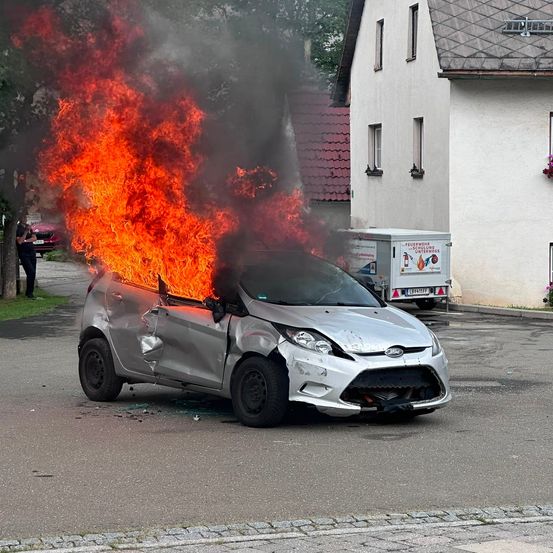 Ein silberner Ford Fiesta steht in Flammen auf einer Straße, mit einem geparkten Feuerwehrwagen in der Nähe. Die Front des Autos ist schwer beschädigt.