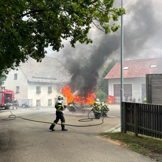 Feuerwehrleute bekämpfen einen Fahrzeugbrand vor einem Haus, wobei Rauch in die Luft aufsteigt.
