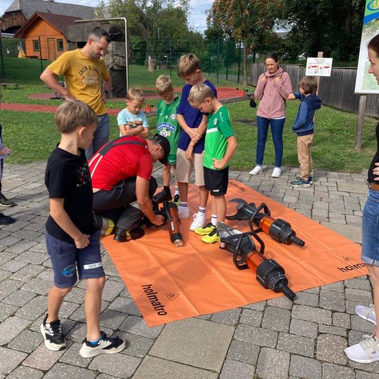Eine Gruppe von Kindern und Erwachsenen ist um eine orangefarbene Plane mit Werkzeugen darauf versammelt, möglicherweise für eine Demonstration oder eine Bildungsaktivität.