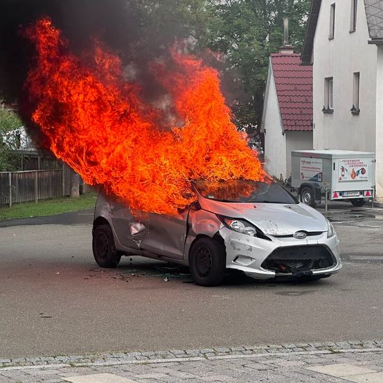 Ein silberner Wagen steht in einem Parkplatz in Flammen, mit einem Anhänger für Feuerlöscher in der Nähe.