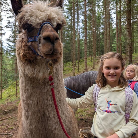 Ein junges Mädchen lächelt, während es neben einem braunen Alpaka im Wald steht. Eine weitere Mädchen ist im Hintergrund.