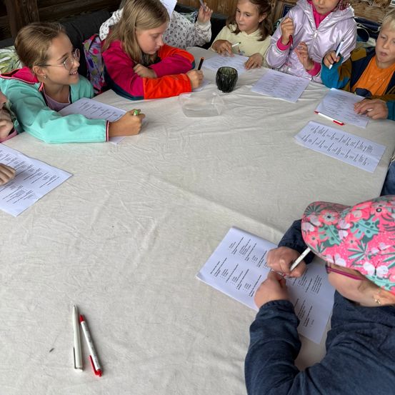 Eine Gruppe von Kindern und einem Erwachsenen sitzen um einen Tisch herum und sind in einer Aktivität vertieft. Sie halten Stifte und schreiben auf Papiere. Der Tisch ist mit einem weißen Tuch bedeckt.