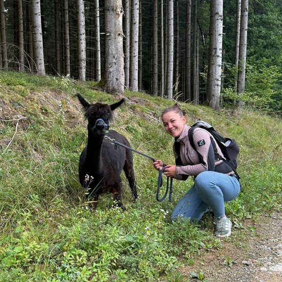 Eine Frau kniet auf einem Grashang, lächelt und hält eine Leine mit einem schwarzen Alpaka neben ihr. Hohe Bäume umgeben das Gebiet.