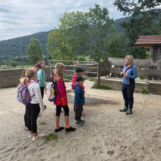 Eine Frau hält eine Rede vor einer Gruppe von Kindern in einem Bauernhof, mit einem Zaun und Bäumen im Hintergrund.