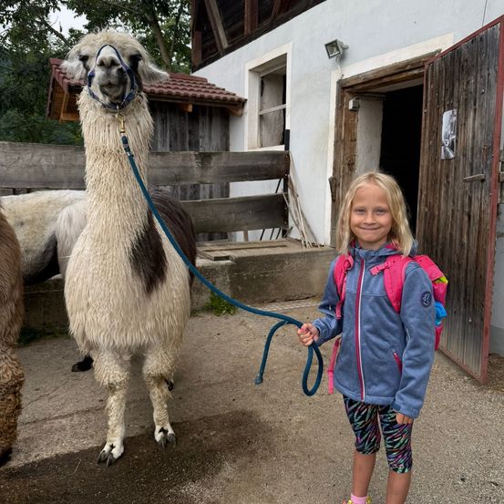 Ein junges Mädchen mit Rucksack steht lächelnd neben einem Alpaka vor einem Holzstall.