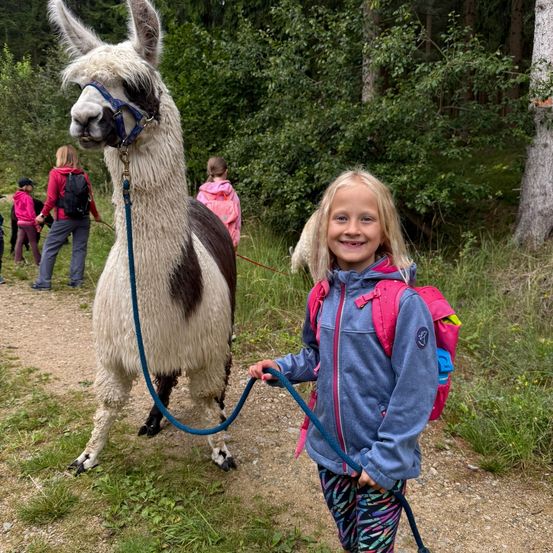 Ein junges Mädchen mit Rucksack geht neben einem lächelnden Alpaka auf einem Grasweg im Wald.