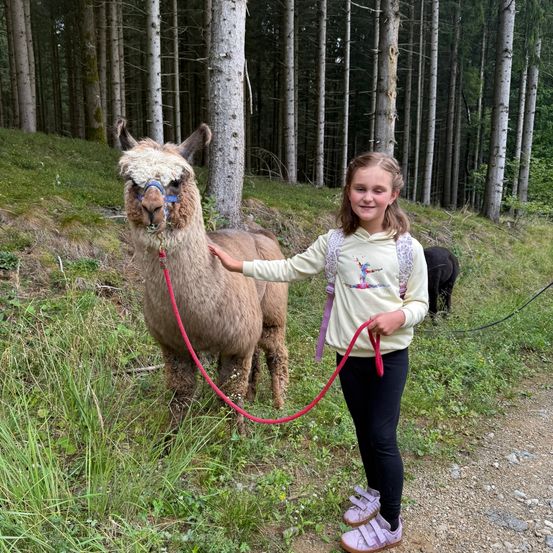 Ein junges Mädchen steht neben einem braunen Alpaka auf einem Grasweg im Wald. Sie hält eine rote Leine, und ein schwarzer Hund ist im Hintergrund zu sehen.