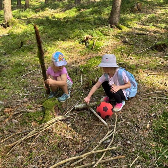 Zwei kleine Mädchen spielen im Wald, eine hält einen Stock und die andere zeigt auf einen roten Fußball.