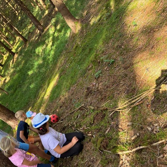 Eine Gruppe von Kindern sitzt auf dem Boden in einem Wald, einige tragen Hüte und einer hält einen Fußball.