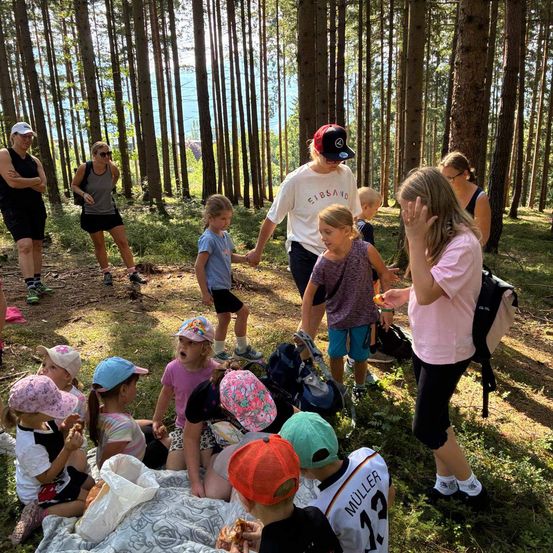 Eine Gruppe von Kindern und Erwachsenen versammelt sich in einem Wald, einige sitzen auf einer Decke, während andere stehen. Ein Erwachsener hält die Hand eines Kindes, und ein anderes Kind isst einen Snack.