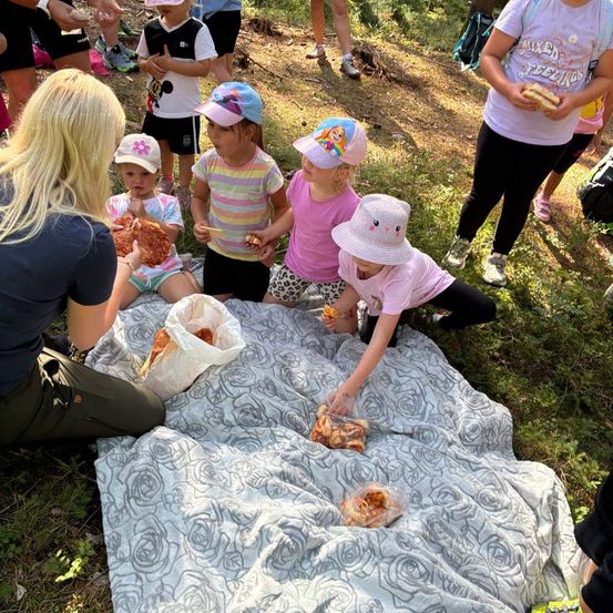 Eine Gruppe von Kindern und Erwachsenen versammelt sich auf einem Grasfeld, einige essen von einem Picknicktisch. Eine Frau in einem blauen Shirt sitzt auf dem Boden und füttert ein Kind.