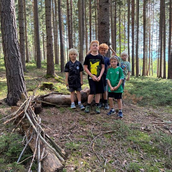 Eine Gruppe junger Jungen steht in einem Wald und lächelt für ein Foto. Sie sind von hohen Bäumen und Grün umgeben.