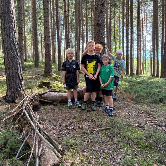 Fünf Kinder stehen in einem Wald, lächeln und posieren für ein Foto. Sie sind von hohen Bäumen und Grün umgeben.