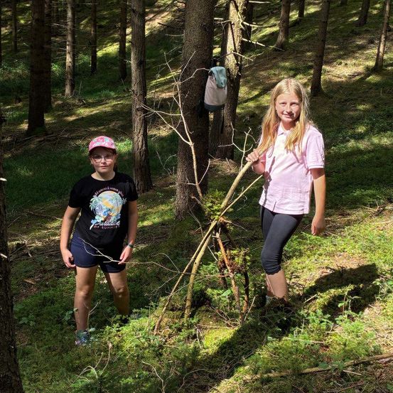 Zwei Mädchen stehen in einem Wald, eine hält einen Stock und die andere lächelt. Das Mädchen links trägt eine Brille und ein schwarzes Shirt. Hinter ihnen hängt an einem Baumstamm eine Tasche.
