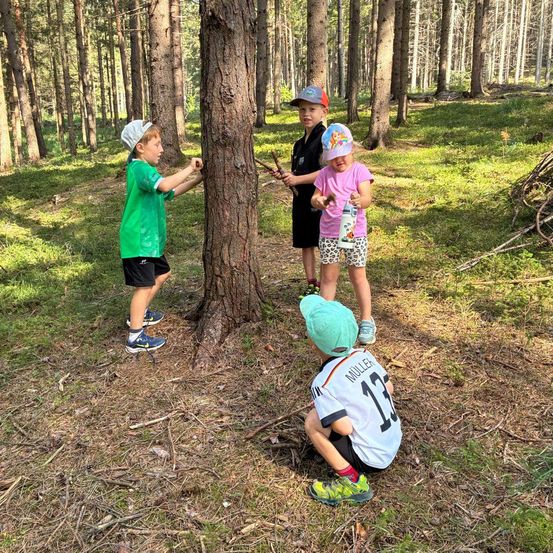 Vier Kinder stehen in einem Wald, wobei ein Junge einen Baumstamm und ein anderes Kind einen Stock hält. Das Mädchen in der Mitte hält eine Flasche. Der Junge vorne kniet auf dem Boden.
