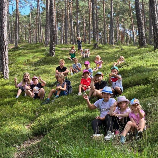 Eine Gruppe von Kindern und Erwachsenen sitzt auf einem Grashügel in einem Wald, einige tragen Hüte und Rucksäcke. Sie lächeln und posieren für ein Foto.