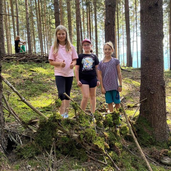 Drei Mädchen stehen in einem Wald und posieren für ein Foto. Das Mädchen auf der linken Seite gibt einen Daumen hoch.