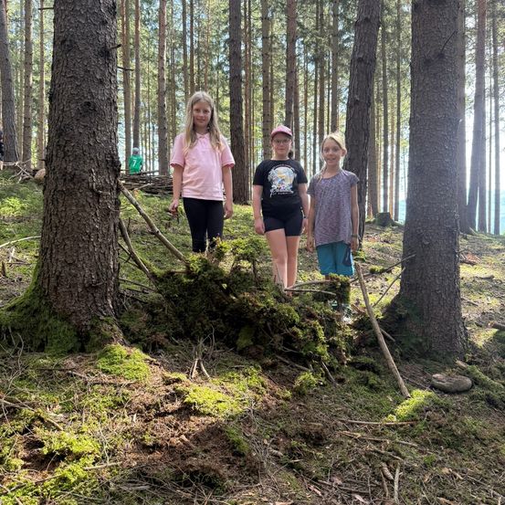 Drei Mädchen stehen in einem Wald und lächeln für ein Foto. Das Mädchen auf der linken Seite hält einen Stock. Das Mädchen in der Mitte trägt eine Mütze und eine Brille. Das Mädchen auf der rechten Seite trägt ein Armband.