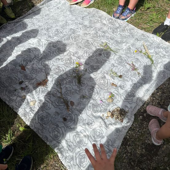 Eine Gruppe von Menschen sitzt auf einer Blumendecke mit verschiedenen Pflanzen und Steinen darauf. Einige von ihnen tragen Turnschuhe.