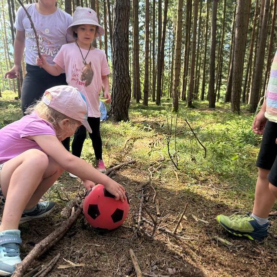 Drei junge Mädchen im Wald, eine kniet und hält einen roten Fußball, während eine andere einen Stock hält und eine dritte hinter ihnen steht.