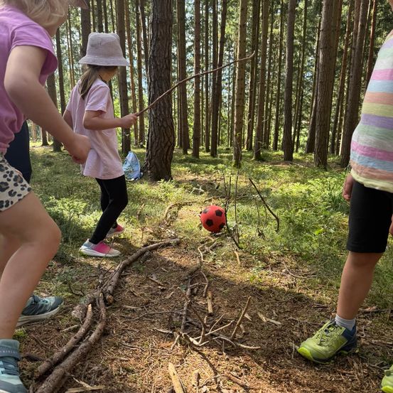 Drei Kinder im Wald, eines hält einen Stock, ein anderes einen Fußball und ein drittes läuft.