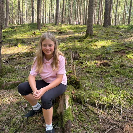 Ein junges Mädchen mit blonden Haaren sitzt auf einem Baumstumpf im Wald, trägt ein rosa Shirt und schwarze Hose. Der Waldboden ist mit Moos und kleinen Pflanzen bedeckt.