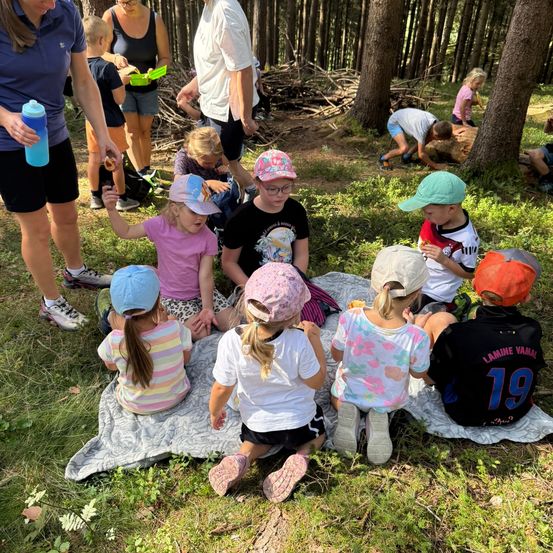 Eine Gruppe von Kindern und Erwachsenen sitzt auf einer Decke im Wald, einige essen, während andere am Boden spielen.