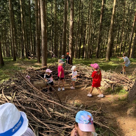 Eine Gruppe von Kindern spielt im Wald, einige bauen ein Nest aus Zweigen. Sie tragen Mützen und Turnschuhe.