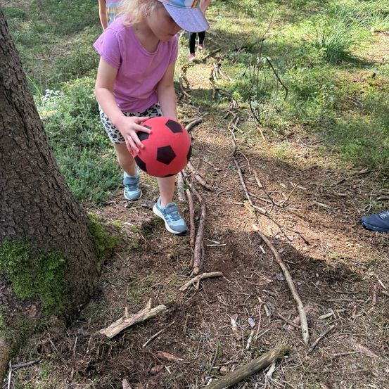 Ein kleines Mädchen hält einen roten Fußball mit schwarzen Akzenten in einem Grasbereich mit einem Baumstamm in der Nähe.