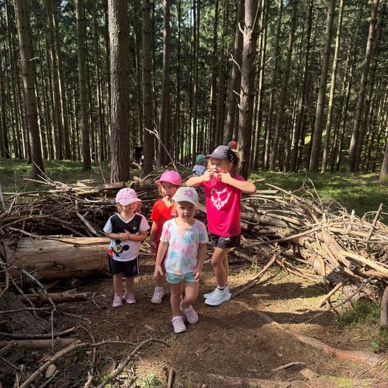 Vier junge Mädchen stehen in einem Wald, zwei vorne und zwei hinten, mit einem Haufen Stöcke im Vordergrund. Sie tragen Mützen und Turnschuhe.