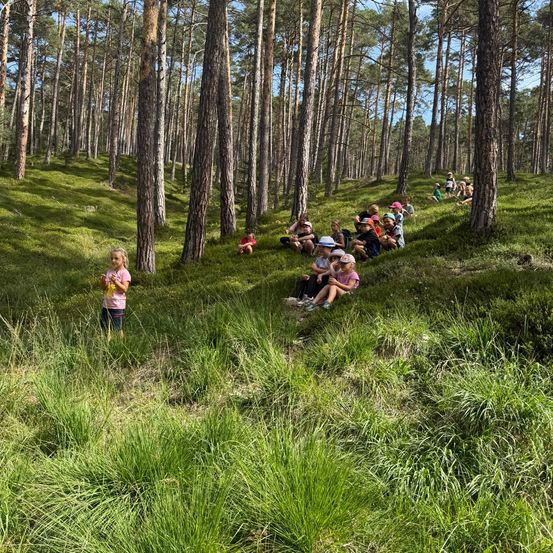 Eine Gruppe von Kindern und Erwachsenen sitzt und steht in einem Wald, einige tragen Hüte und Schuhe. Ein Mädchen steht im Vordergrund.